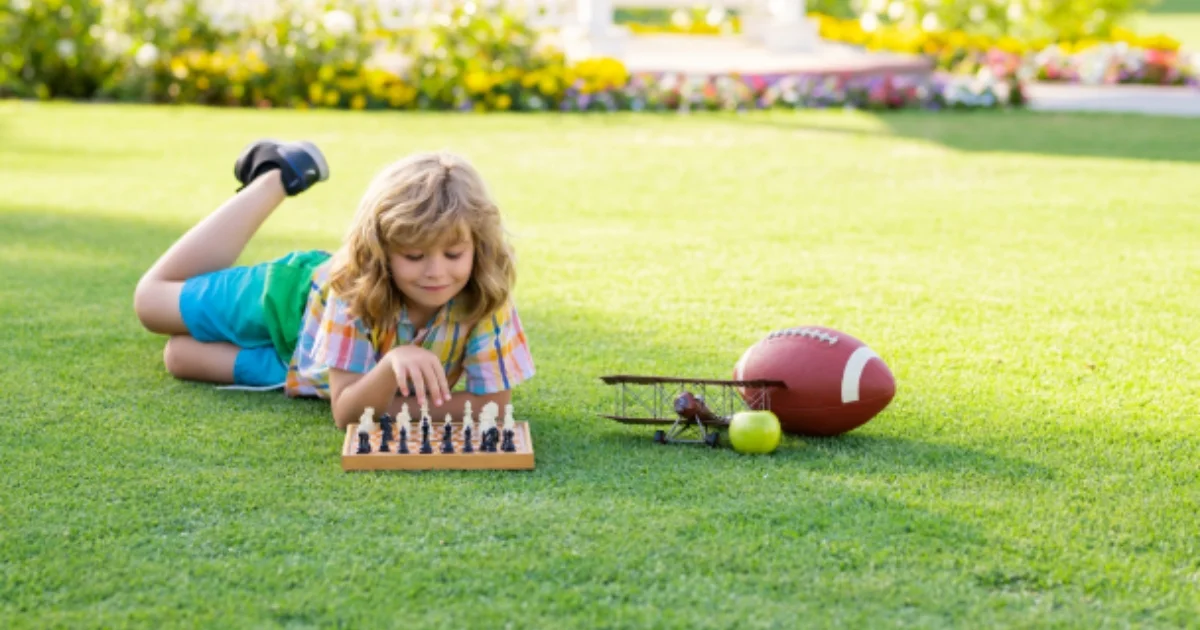 Young child playing chess on a lawn with toys and a football beside them.